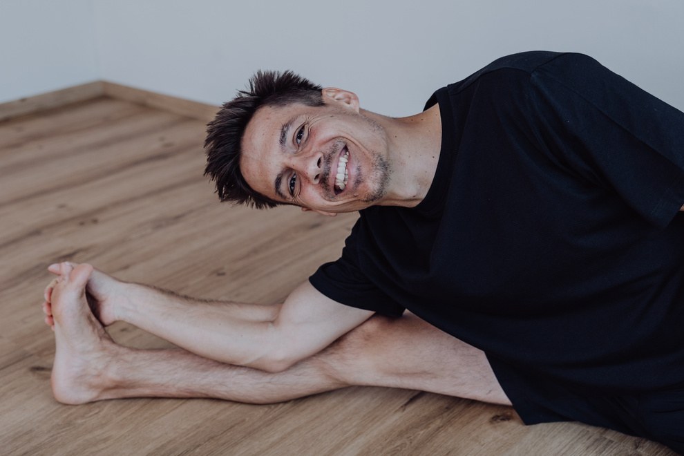 Manuel sitzt in der Vorbeuge auf dem Studioboden, hält seine Füße und lacht in die Kamera